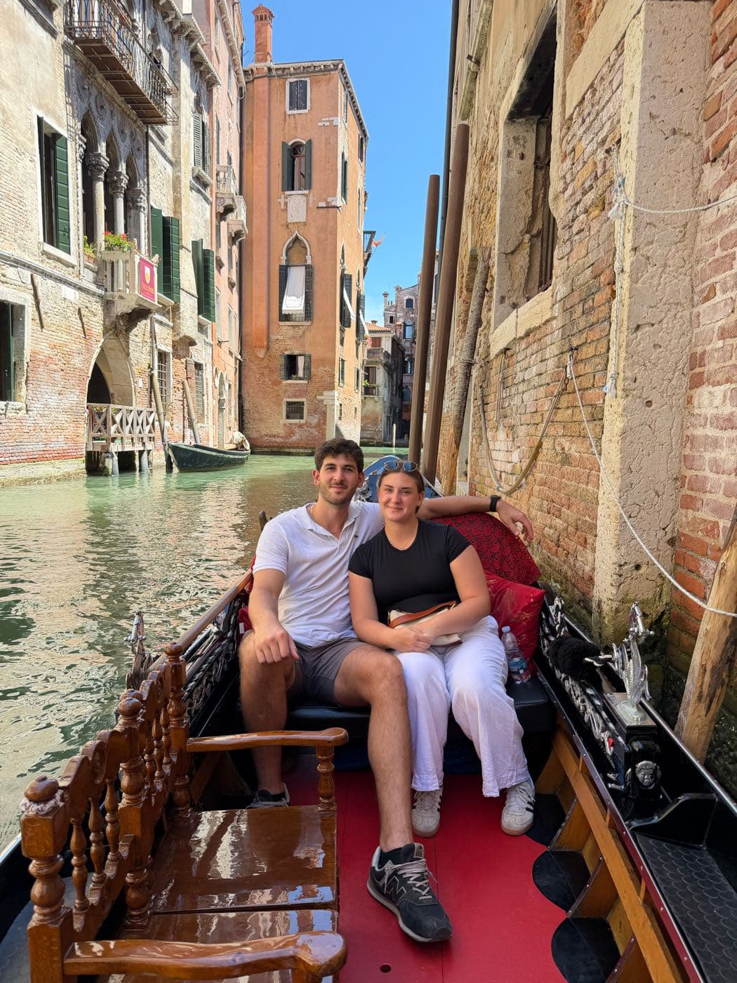 Lisa and Marko sitting close together in a gondola on a canal in Venice, old brick buildings on either side